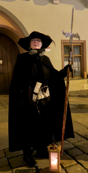 Die Nachtwächterin von Landshut in schwarzem Umhang mit breitkrempigen Hut steht nachts vor dem Rathaus in der Altstadt von Landshut. In ihrem Gürtel steckt ein Signalhorn und sie hält eine Hellebarde in der Hand.